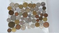 Photo showing approximately 57 assorted coins scattered on a white surface, with various sizes and colors, some copper, some silver, some gold toned.