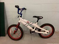 Front-side view of the white Pacific Cycle NHL bicycle with 15-inch wheels, blue handlebars, black seat with hockey graphics, and red rims with decorative spokes.