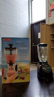 Wide shot showing Back to Basics smoothie maker next to its original box and Osterizer blender in background, placed on a dark surface near a window.