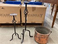 Photo showing two wrought iron candle holders of different heights and a vintage copper pot with handles on concrete floor with boxes in the background.