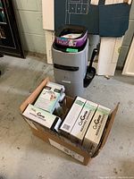 Photo showing the Cat Genie litter disposal system unit with purple lid and handle, alongside an open cardboard box filled with several sealed boxes of Cat Genie washable granules and supplies.