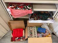 Four cardboard boxes containing various Christmas decorations including ornaments, mini string lights, and fabric items like a table cloth.