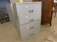 Four drawer metal filing cabinet with label holders and recessed handles on each drawer. Cabinet is light grey and shows slight signs of use.