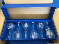 Six clear glass goblets arranged in a row inside a blue box with individual slots. The box interior is tan with writing on the inside of the lid.
