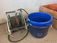 Green garden hose coiled on white and green plastic reel stand next to two plastic buckets on concrete floor near cinder block wall