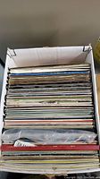 Image of a cardboard box filled with approximately 40-50 vinyl LP records stacked vertically, showing a side view of many record sleeves.