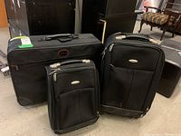 Three pieces of black luggage with one large American Flyer suitcase and two Samsonite suitcases in front of a piano and furniture.