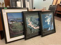 Three framed prints with black frames leaning against a wall, showing two golf course images and one snowboarding image.