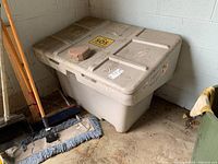 Photo of a used off-white plastic salt/sand bin placed against the wall, showing the rectangular storage container with hinged lid and handles, with a brick on top and cleaning tools nearby.