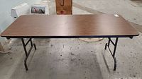 Rectangular folding table with faux wood top and metal legs in warehouse setting, showing top surface and legs in fully set up position