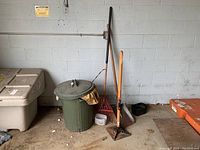 Photo showing green plastic garbage can with lid, red metal rake with wooden handle, metal tamper with wooden handle, and small shovel with wooden handle against gray cinderblock wall