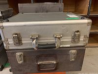 Front view of a silver metal case stacked on top of a brown faux leather case, showing latch locks and handle details.