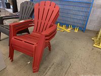 Three red resin stackable outdoor Adirondack style chairs with slatted back and armrests, photographed indoors on concrete floor.