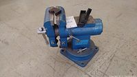 Larin blue cast metal 5 inch bench vise on the floor showing the clamping jaws, rotating base, and a mounted handle.
