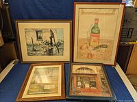 Photo showing all four framed art pieces laid out on a blue cloth; includes Dubunet painting, watercolor still life, Missouri poster, and Antoinette storefront print.