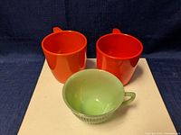 Two red Rosti Denmark cups and one green Fire King cup, shown on a white surface with dark blue background.