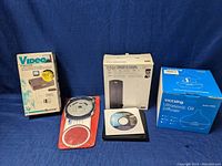 Full lot photo showing all five boxed and loose items on a blue fabric background: Recoton RF Modulator box, CPC Flight Computer sealed in plastic, Terk Edge Powered FM Antenna box, Samsung Blu-ray Writer with installation disc in front, VicTsing Ultrasonic Oil Diffuser box.