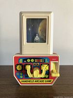Front view showing the game's display featuring a spaceship in space with moon background and control panel with yellow knobs and lever.