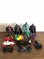 Four Batman figures, Robin, Joker and Penguin action figures, and multiple toy vehicles including Batmobiles and cartoon-style cars on a wooden surface.