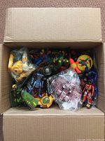 Overhead photo showing various mixed action figures and toys inside an open cardboard box including a yellow duck, a blue armored figure, and some wrapped plastic items.