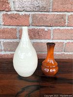 Photo of both decorative vases side by side with brick background and wooden surface, showing size comparison and surface detail.