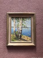 Full view of the framed painting showing birch trees by a lake with a wood frame on a carpeted floor.