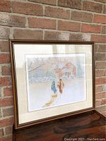 Framed print showing two people walking on a snow-covered path towards a house in the background, with bare winter trees around, in a brown wooden frame with double matting.