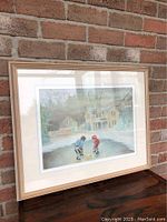 Framed print depicting two children playing hockey outside in front of a house during winter, in a light wood frame with double matting under glass.