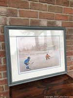 Full frontal photo of the framed print showing the winter hockey scene with two players on an iced pond, staged against a brick backdrop.