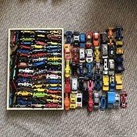 Photo showing large wooden tray filled with multiple rows of assorted diecast toy cars including classic and racecars in various colors and designs