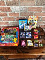 Wide shot of all Pokémon items laid out on wooden table against brick wall showing sticker kits, card albums, booster packs, collectibles, and loose cards.