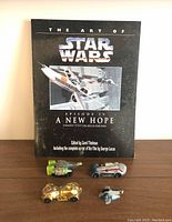 Hardcover art book The Art of Star Wars Episode IV A New Hope 1995 with four Star Wars-themed vehicles placed on a wooden surface.