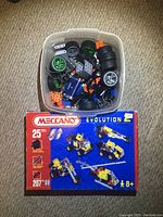 Plastic tub filled with mixed Meccano parts including wheels and metal plates, with Meccano Evolution 2 box