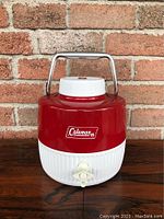 Front view of red and white Coleman 1 gallon water jug with metal handle and spigot.