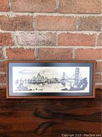 Framed art print depicting San Francisco cityscape with Golden Gate Bridge against a brick background on a wooden surface.
