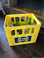 Yellow Pepsi plastic delivery crate with mixed glass soda bottles inside viewed at an angle showing front and left sides of the crate and bottles inside.