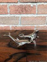 Pair of silver plated animal shaped bottle openers, one seal and one dolphin, shown from a side angle against a brick background on a wooden surface.