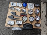 Photo showing 18 various car lights laid out on brown cardboard including fog lights, headlights, marker and signal lights, in various shapes and colors like white, clear, blue, and red.