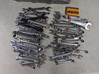 Overview of most of the wrenches laid out on a floor with some stacked, showing various sizes and some brand marks, includes a small plastic storage bin partially visible at the top left.