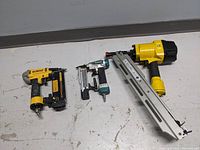 Three air nailers placed side by side on the floor showing size and condition, includes Powerfist framing nailer, DeWalt nailer, and two smaller Makita nailers.
