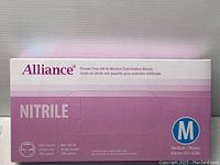 Front of box showing Alliance branding, NITRILE text, medium size indicator (M), and product details including powder free, non sterile, single use, 100 pieces.