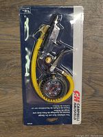 Photo of the Campbell Hausfeld tire inflator in original packaging, showing the inflator gun, yellow hose, and 2-inch black pressure gauge.