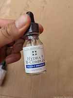 Photo of a hand holding a small 15ml glass bottle with black dropper cap. Label shows Cellex-C Enhancer Hydra 5 B-Complex gel hydratant.