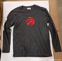 Dark gray long sleeve shirt laid flat showing Raptors claw basketball logo on front center.