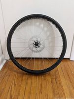 Photo of a 26x4 inch black fat tire rim with disc brake rotor mounted, shown standing upright against a wall on wooden floor.