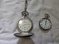 Two pocket watches on a cloth background, one larger with an open cover and chain, the other smaller without cover or chain visible.