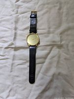 Full view of the men's Komono Winston watch laid flat showing gold-tone round face and black leather band.