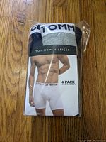 Front image of the packaged 4-pack Tommy Hilfiger boxer briefs showing model wearing white briefs and packaged boxer briefs colors (black, white, gray).
