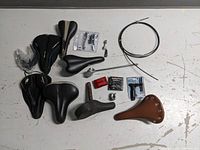 Photo showing several black cushioned bike seats with stems, seat cushions, coiled cables, brake parts in packaging, bike lights and small accessories arranged on floor.