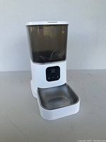 White automatic pet feeder with a clear dark food container and stainless steel feeding bowl shown against a plain background.